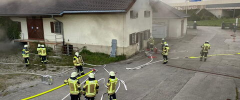 Feuerwehrleute bekämpfen Rauchentwicklung aus einem Gebäude, während Schläuche auf dem Boden ausgerollt sind.
