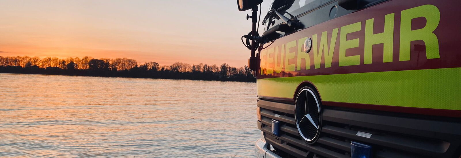 Ein Feuerwehrwagen steht im Sonnenuntergang am Seeufer und spiegelt den Frühlingsabend wider.